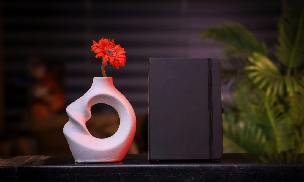 نوت بوك أسود بجانب مزهرية بيضاء وزهرة حمراء، هدايا دعائية فاخرة.