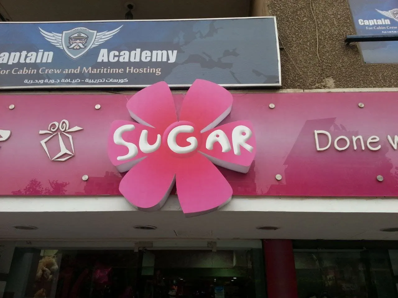 لافتة متجر "SUGAR" وردية لامعة وشعار زهرة، تحت لافتة "Captain Academy" الزرقاء.