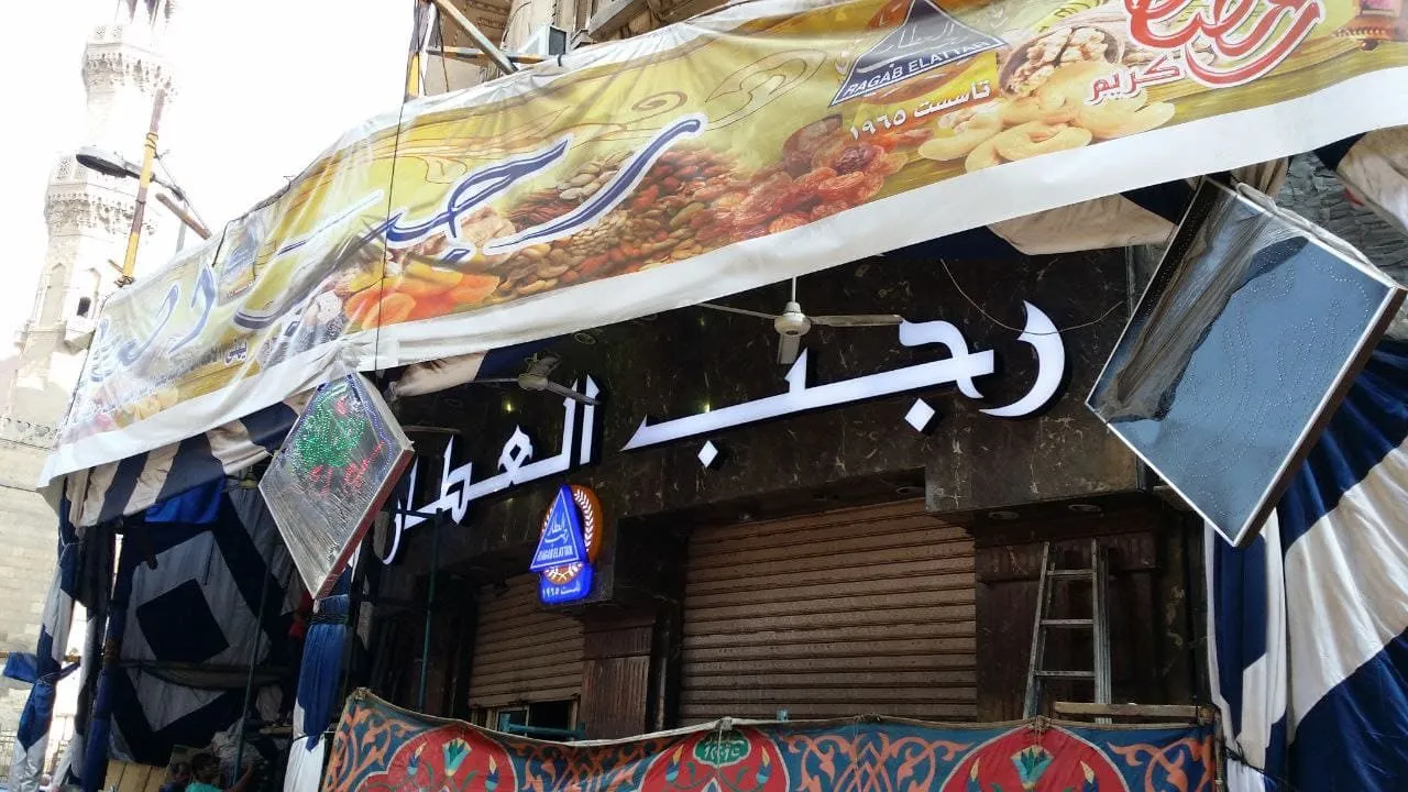 واجهة محل "رجب العطار" للمكسرات والحلويات القديم، لافتة صفراء مضيئة، وأبواب مغلقة.