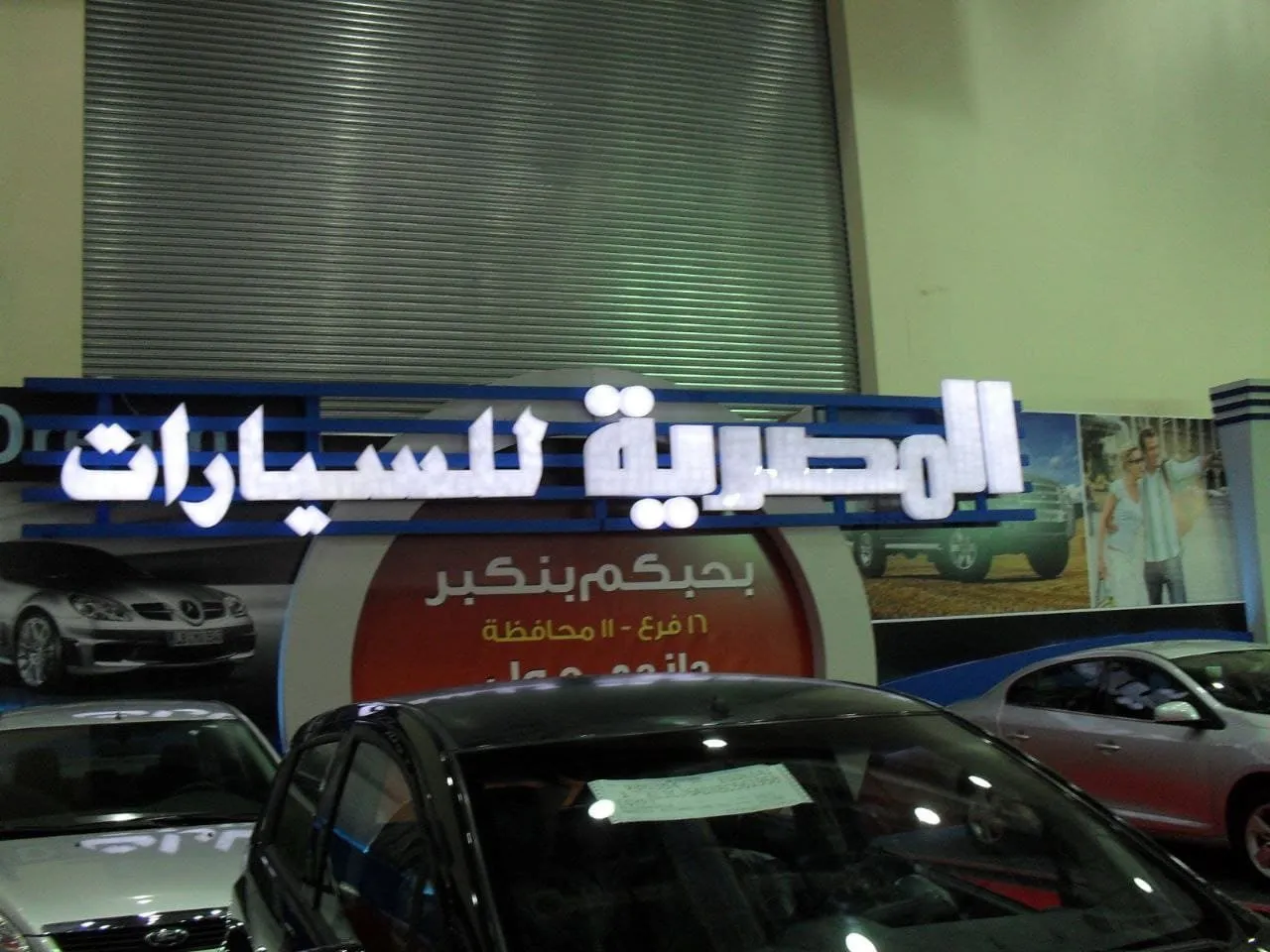 معرض "المصرية للسيارات" يعرض سيارات جديدة وسوداء وفضية أمام لافتة مضيئة.