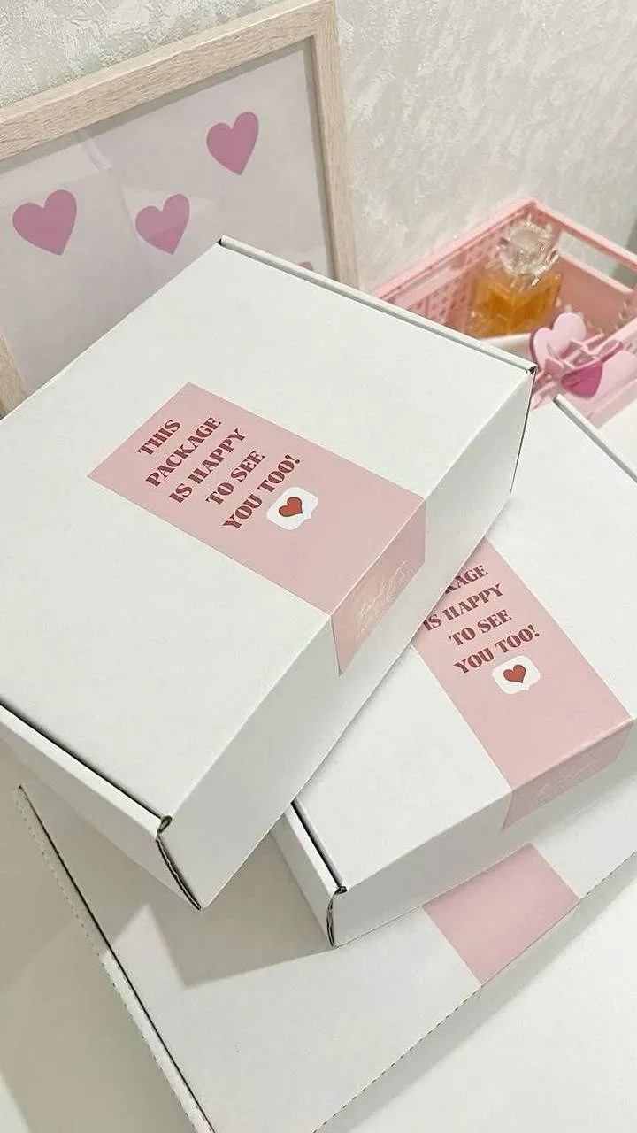 صناديق شحن بيضاء مكدسة عليها ملصق وردي مرح مع زجاجة عطر وديكور قلوب.