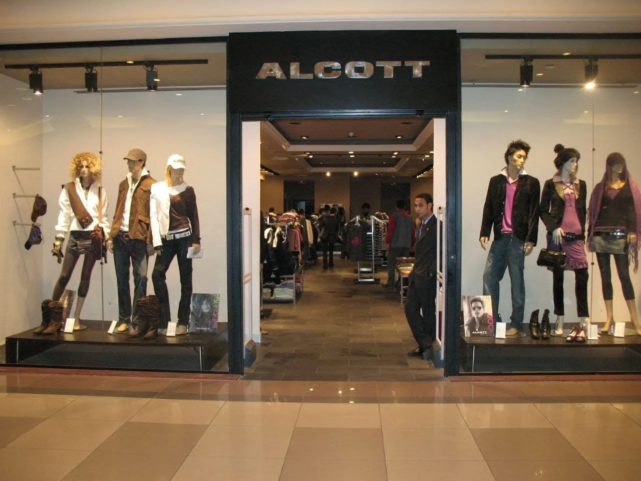 واجهة متجر أزياء "ALCOTT" تعرض ملابس عصرية على مانيكانات مضاءة بمركز تجاري.