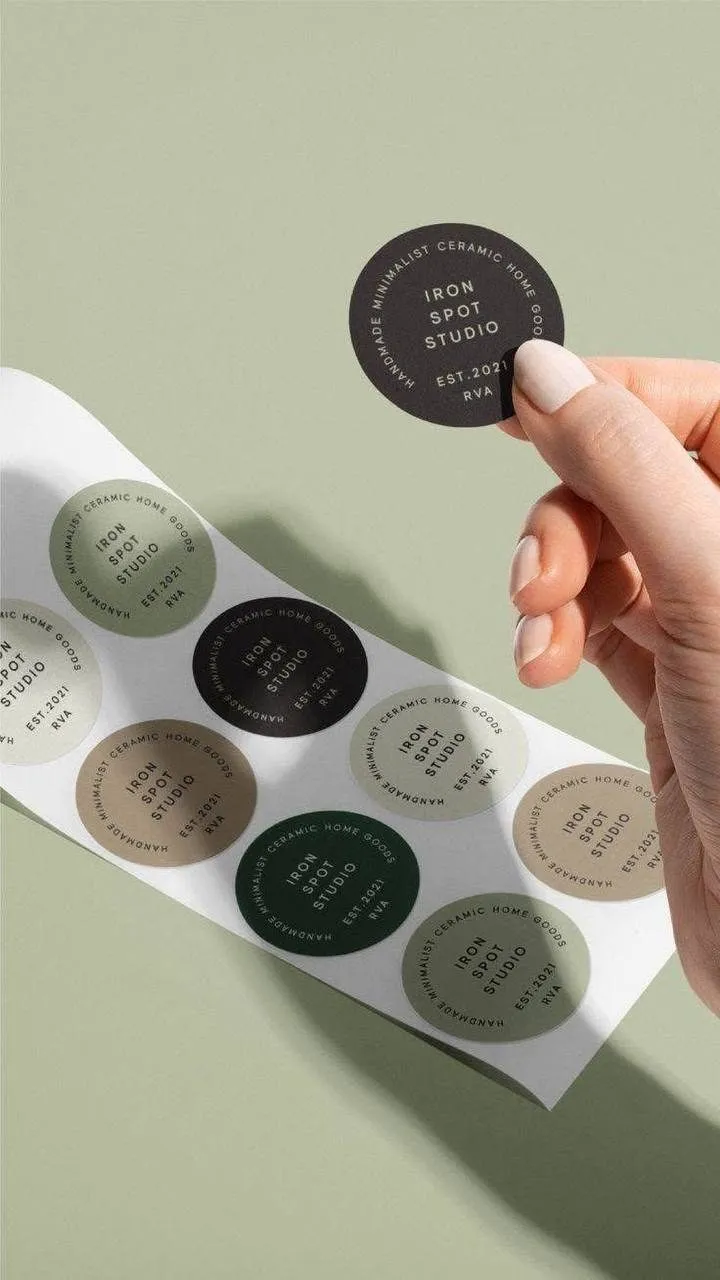 يد تمسك ملصقات دائرية تحمل شعار IRON SPOT STUDIO بألوان ترابية هادئة.
