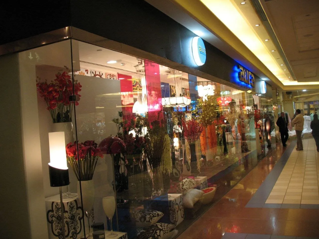 واجهة متجر زجاجية تعرض زهورًا اصطناعية وإكسسوارات منزلية في ممر تسوق مضاء.