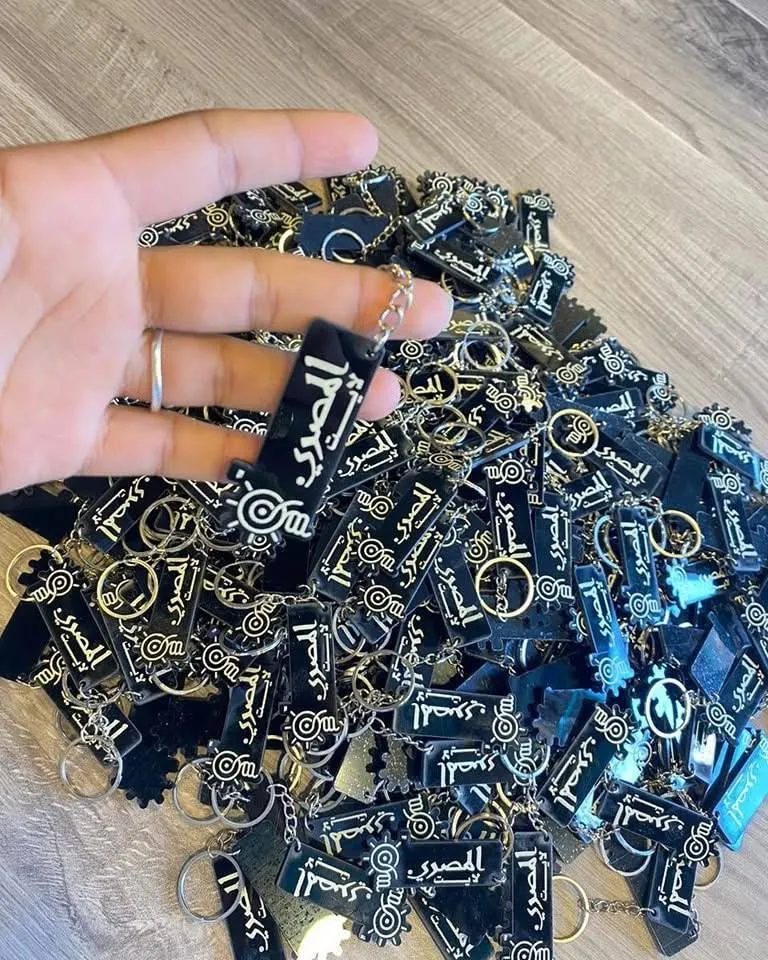 يد تمسك سلسلة مفاتيح سوداء تحمل شعار "المصري" فوق كومة مفاتيح متشابهة.