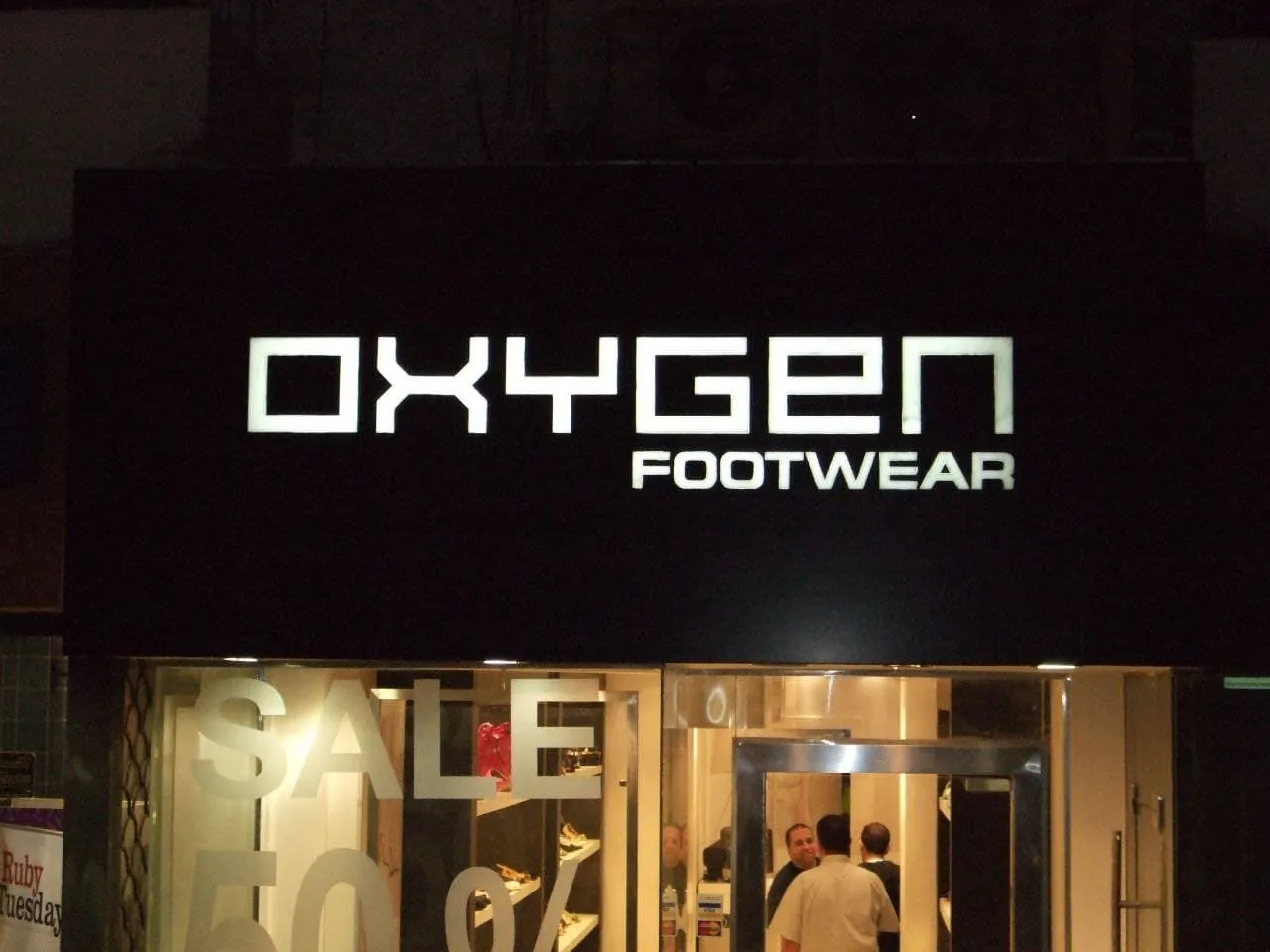 واجهة متجر أحذية "OXYGEN FOOTWEAR" ليلًا مع لافتة مضيئة وإعلان تخفيضات "SALE 50".