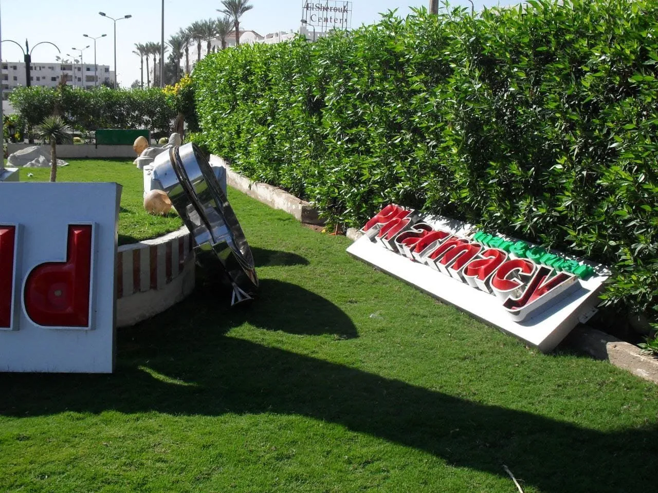 لافتة "Pharmacy" مائلة في حديقة مشمسة مع عشب أخضر وسياج نباتي.