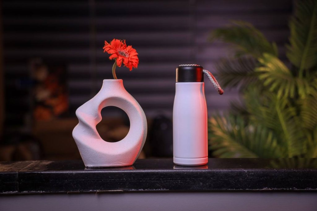 ترمس حراري أبيض أنيق بجوار مزهرية بيضاء وزهرة برتقالية على سطح لامع.